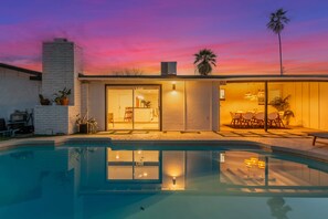 Una casa moderna con piscina, un patio con mobiliario exterior y un cielo al atardecer.