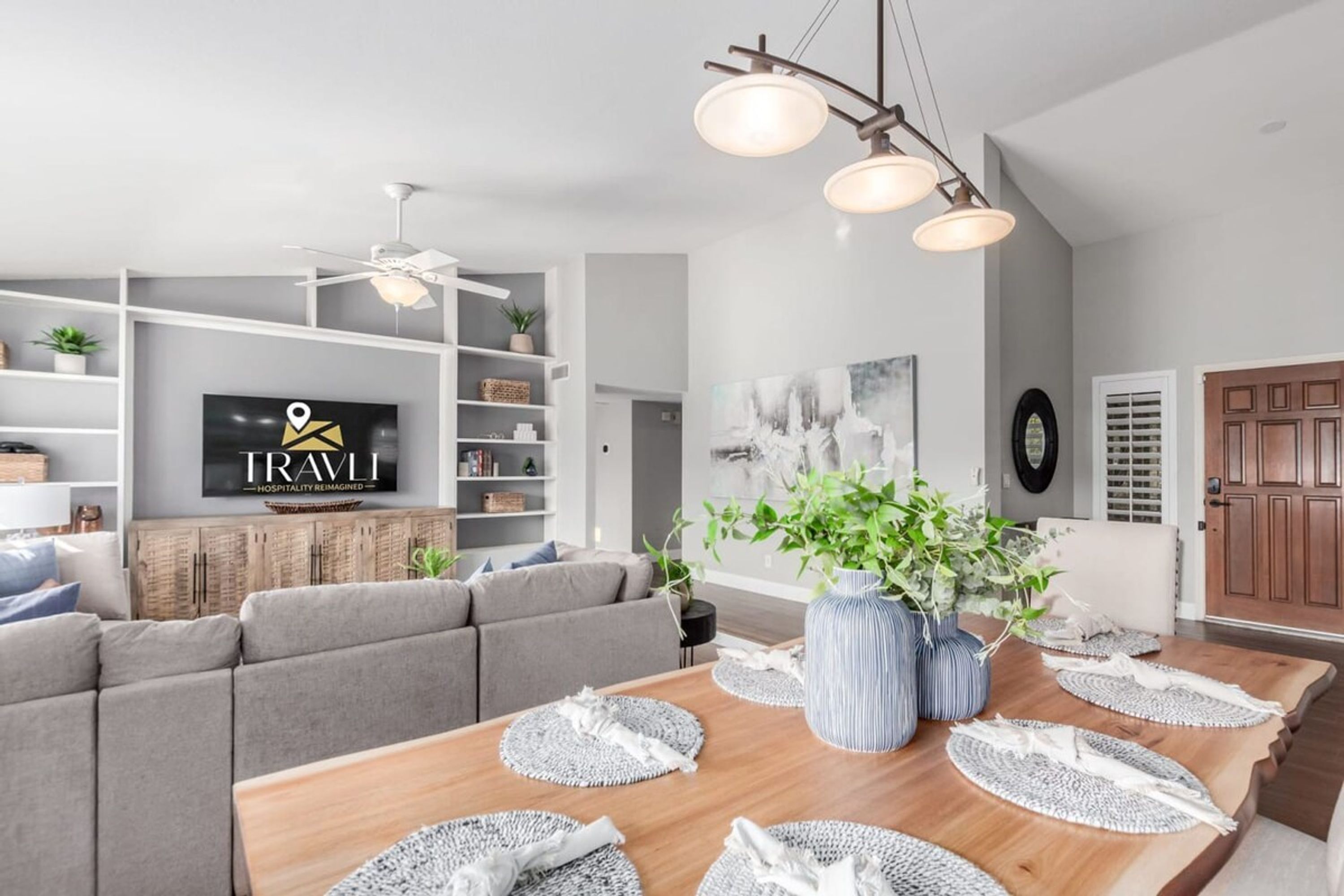Ein modernes Wohnzimmer mit einem Holzesstisch, grauen Sofas und einem Fernseher, der 'TRAVLL' anzeigt.