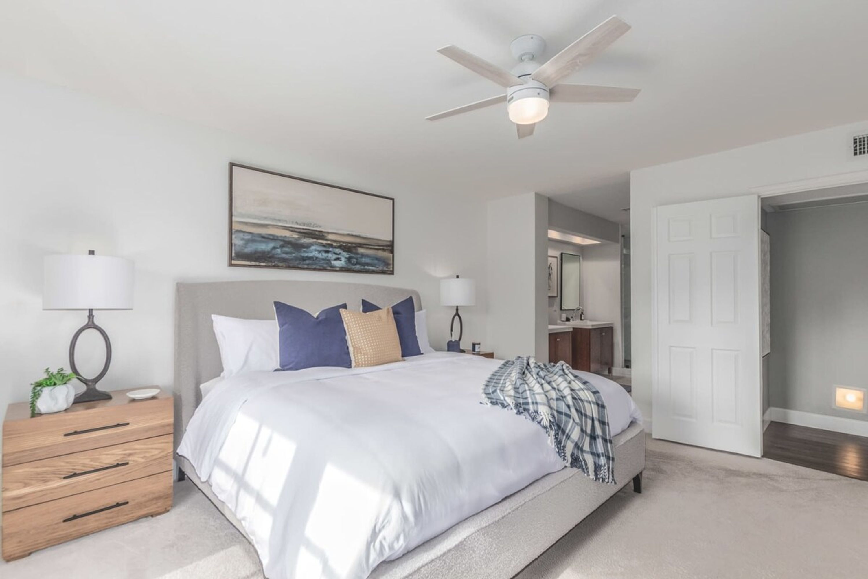 A bedroom with a bed, nightstands, a ceiling fan, a painting, and a door leading to another room.