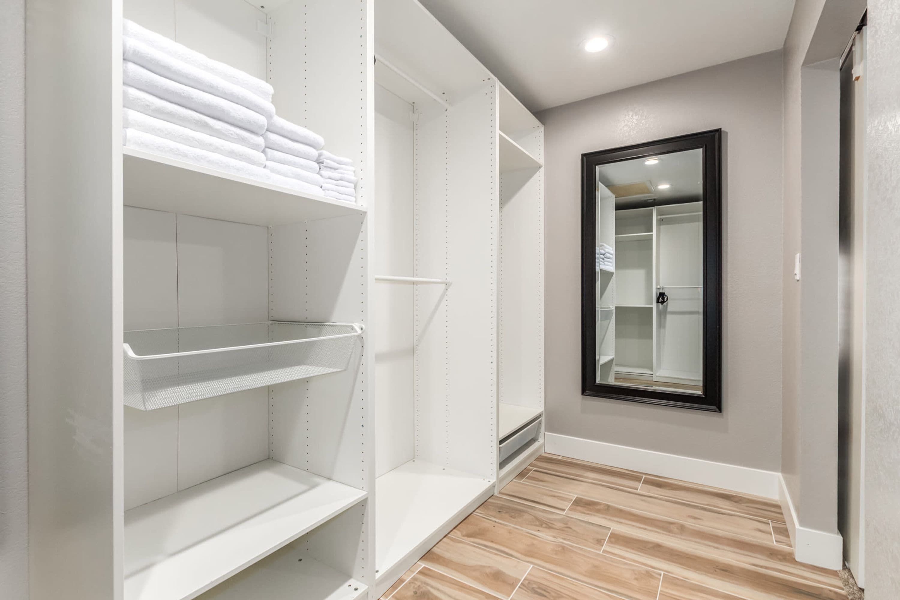 A walk-in closet with white shelving units, a mirror, and a wooden floor.