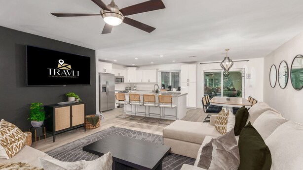 A modern living room with a flat-screen TV, a ceiling fan, and a dining area with a kitchen in the background. - Desert Rose Retreat Heated Pool 2 Masters Gameroom (Scottsdale)