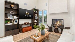 A modern living room with a flat-screen TV, wooden shelving, a fireplace, and a view through the glass door. - Stonewood Luxe Estate 5 Showers Heated Pool GYM (Scottsdale)