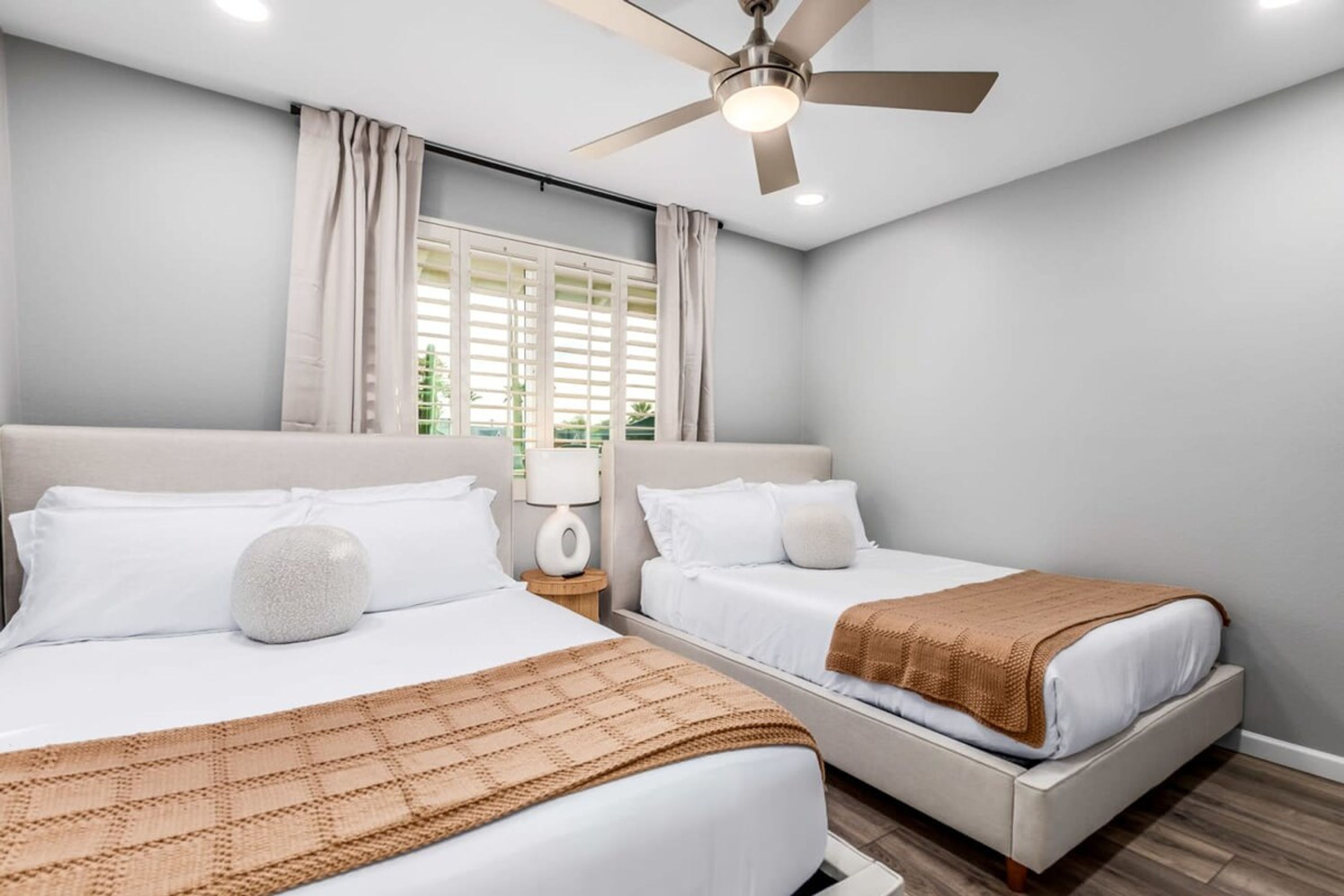 Habitación de hotel con dos camas, un ventilador de techo, una ventana con cortinas y una lámpara en una mesita de noche.