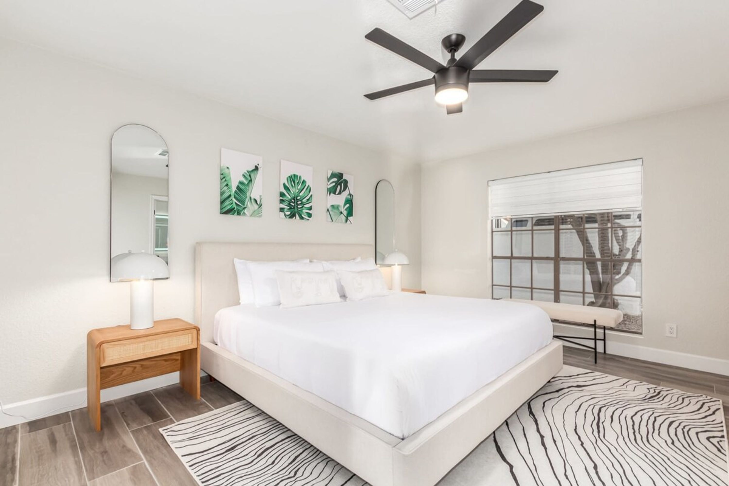 A bedroom with a bed, nightstands, a ceiling fan, a window with a view, and wall art.