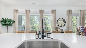 Una cocina con una isla grande, un fregadero y un ventanal con persianas. Hay botellas sobre la isla, un espejo en la pared y una planta a la izquierda.