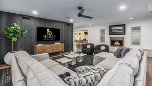 A modern living room with a grey sofa set, a flat-screen TV mounted on the wall, a fireplace, and a dining area in the background.