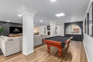 A modern living room with a pool table, a sofa, and a TV displaying the word "TRAVEL". - 8BR Villa Heated Pool Putt Grn Billiards Sleeps 18 (Phoenix)