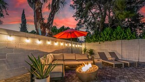 An outdoor patio with a brick wall, a curved sofa, lounge chairs, a fire pit, and string lights.