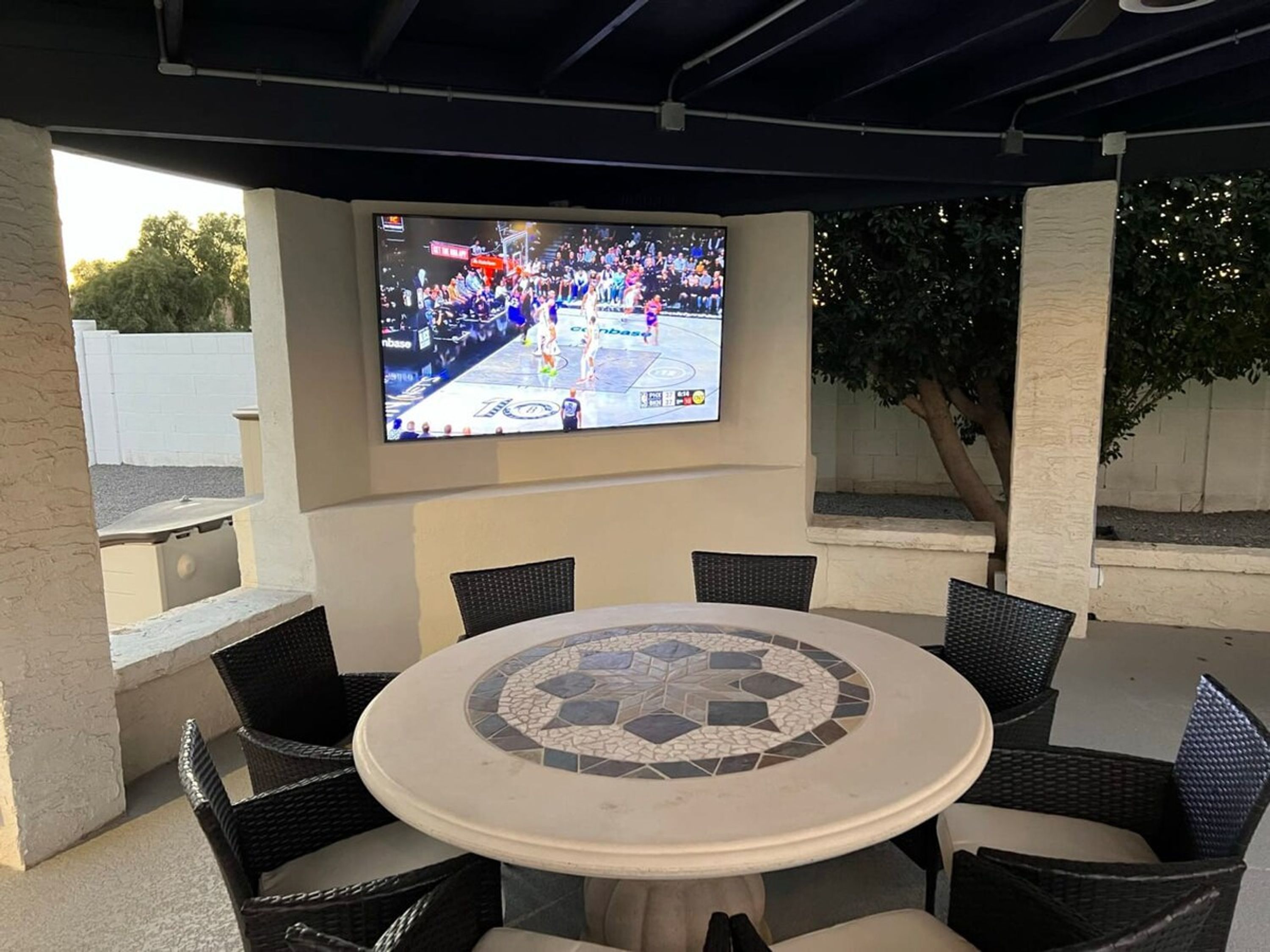 An outdoor patio with a round table and chairs, a flat-screen TV mounted on the wall, and a view of a basketball game.