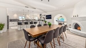 A modern kitchen with white cabinets, a dining area with a wooden table and chairs, a fireplace, and a TV mounted on the wall. - 5BR Resort-style Oasis 2 Primary s Cabana Gameroom (Chandler)