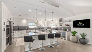 A modern kitchen with white cabinetry, a central island with bar stools, pendant lighting, and a mounted TV. - 5BR Resort-style Oasis 2 Primary s Cabana Gameroom (Chandler)