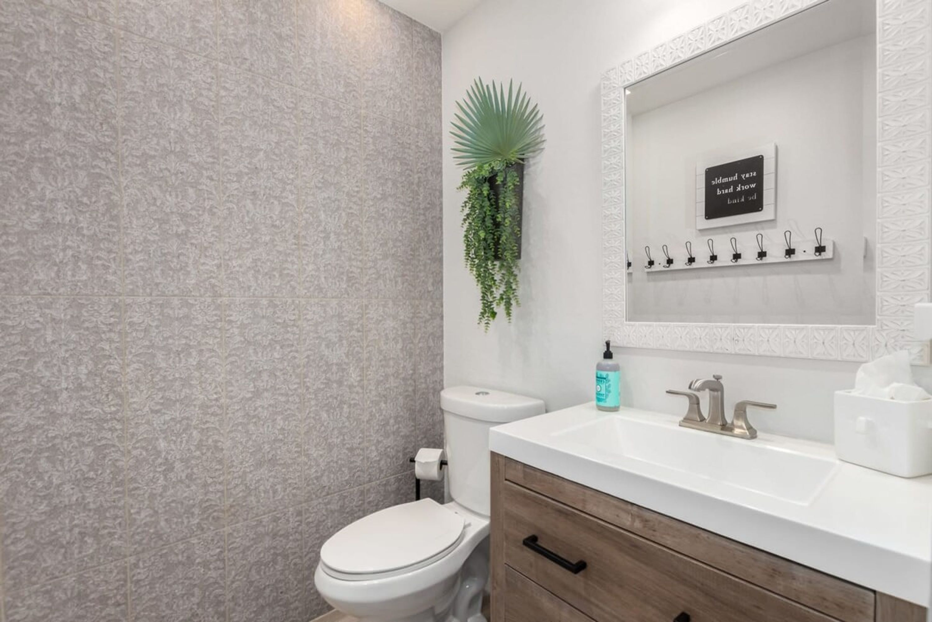 A bathroom with a toilet, sink, and a mirror above it. There is a wall-mounted plant and a sign with text.