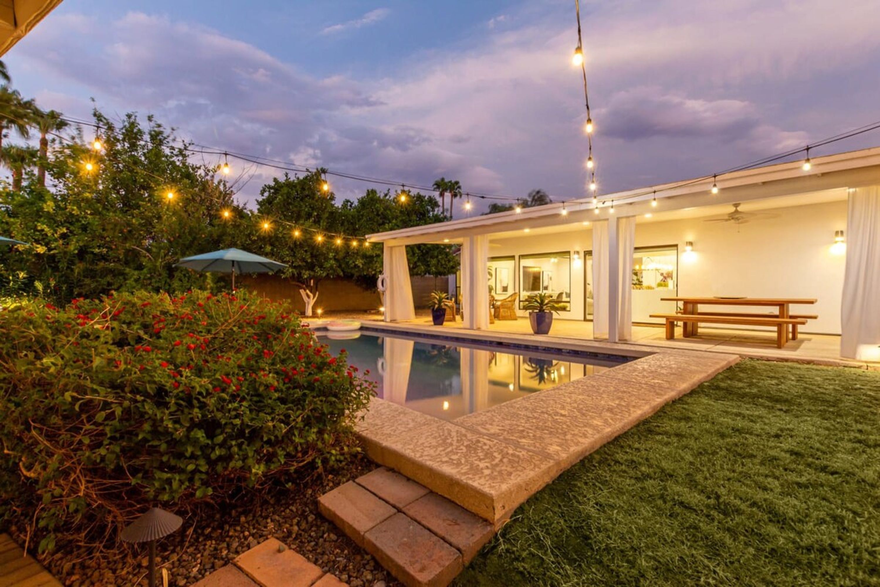 Un patio trasero con piscina, un porche cubierto y luces colgantes.