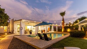 Un patio trasero con piscina, sillas de descanso y luces colgantes.
