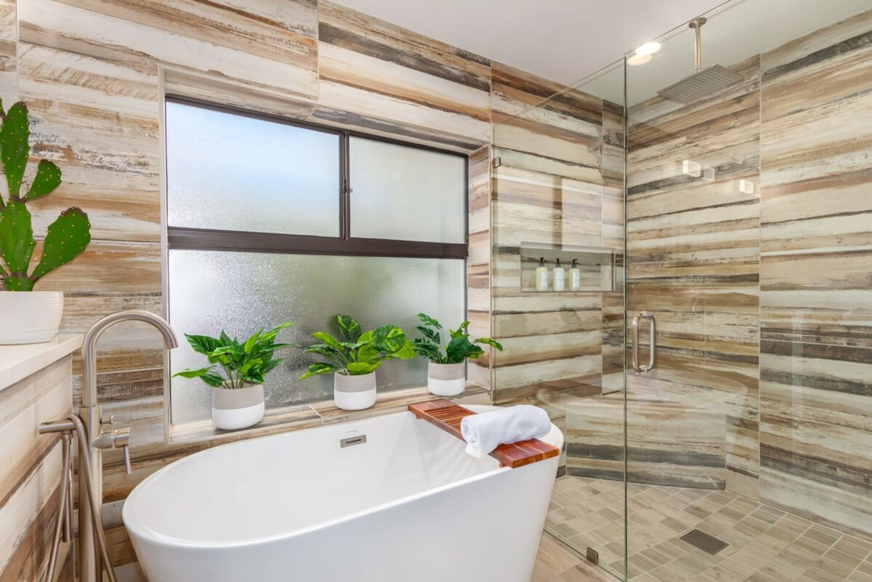 A modern bathroom with a freestanding bathtub, a glass shower enclosure, and three potted plants on the windowsill.