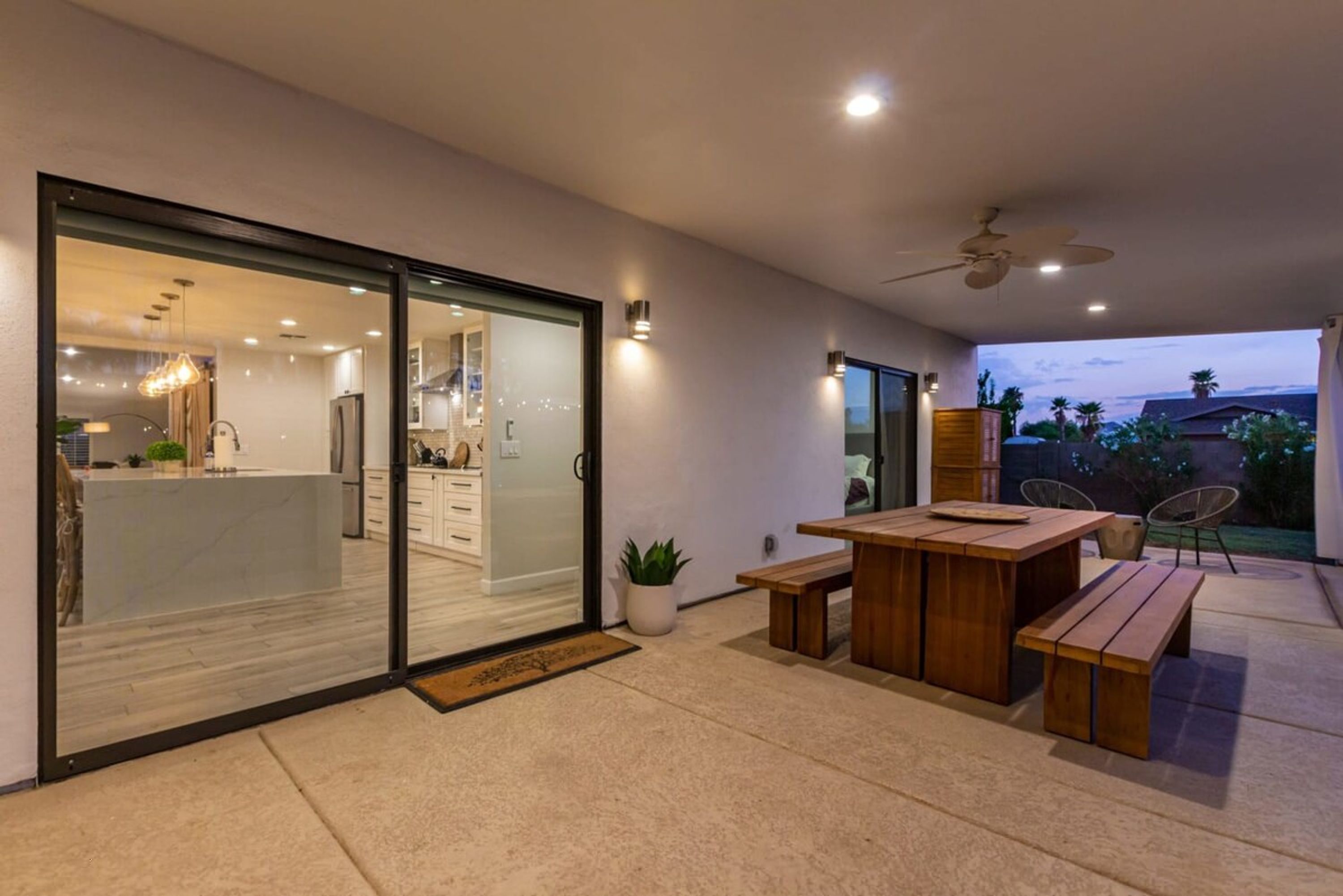A modern patio area with a wooden table and benches, a ceiling fan, and a sliding glass door leading to a kitchen.