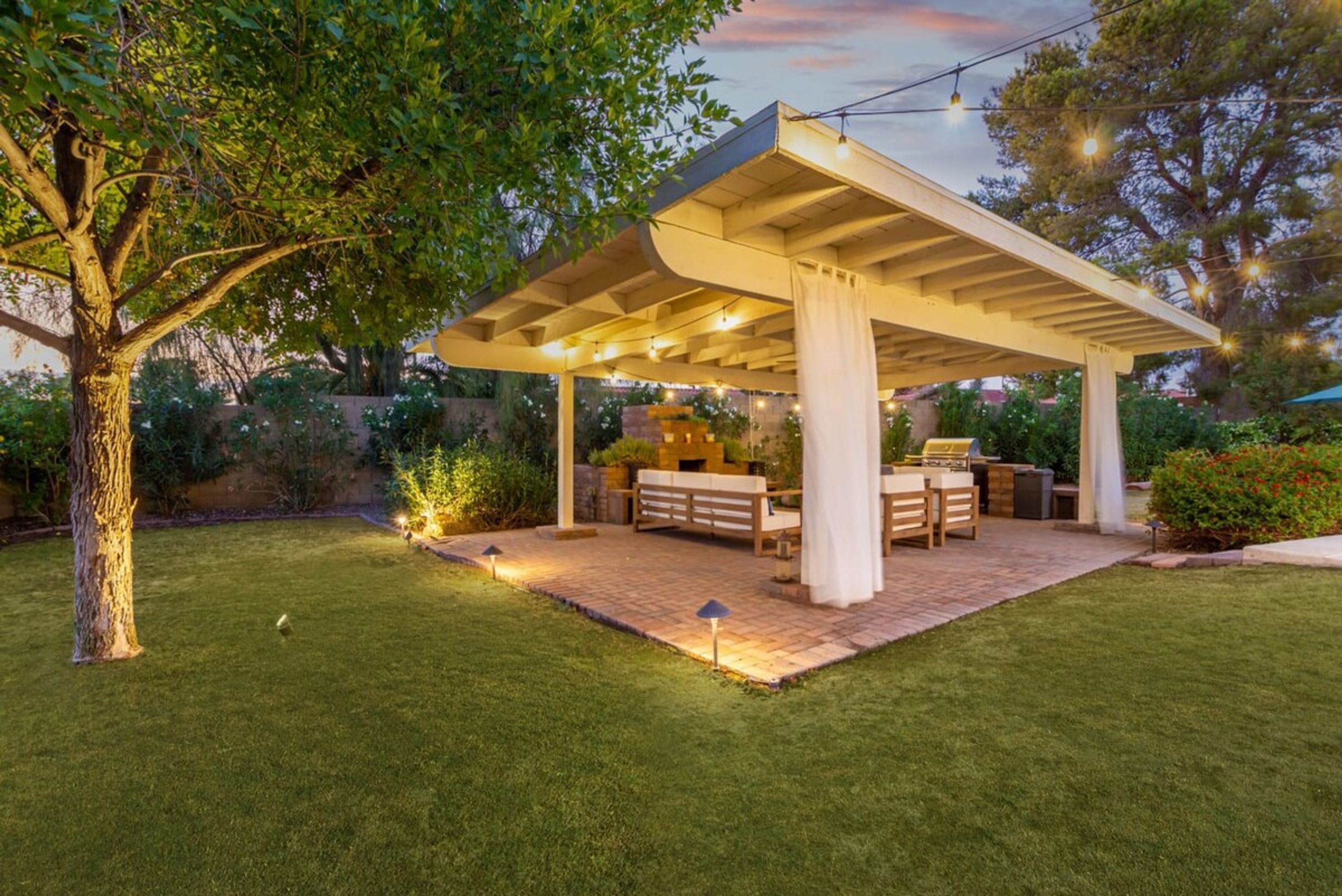 A covered outdoor patio with a dining table and chairs, surrounded by a well-maintained lawn and trees.