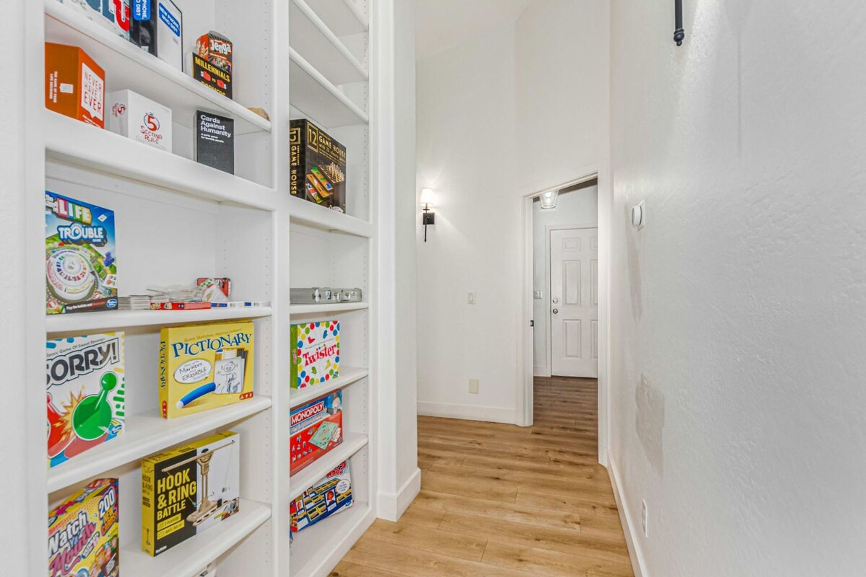 A hallway with white walls and wooden flooring, leading to a door. Shelves on the left hold various board games and puzzles.