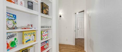 A hallway with white walls and wooden flooring, leading to a door. Shelves on the left hold various board games and puzzles.