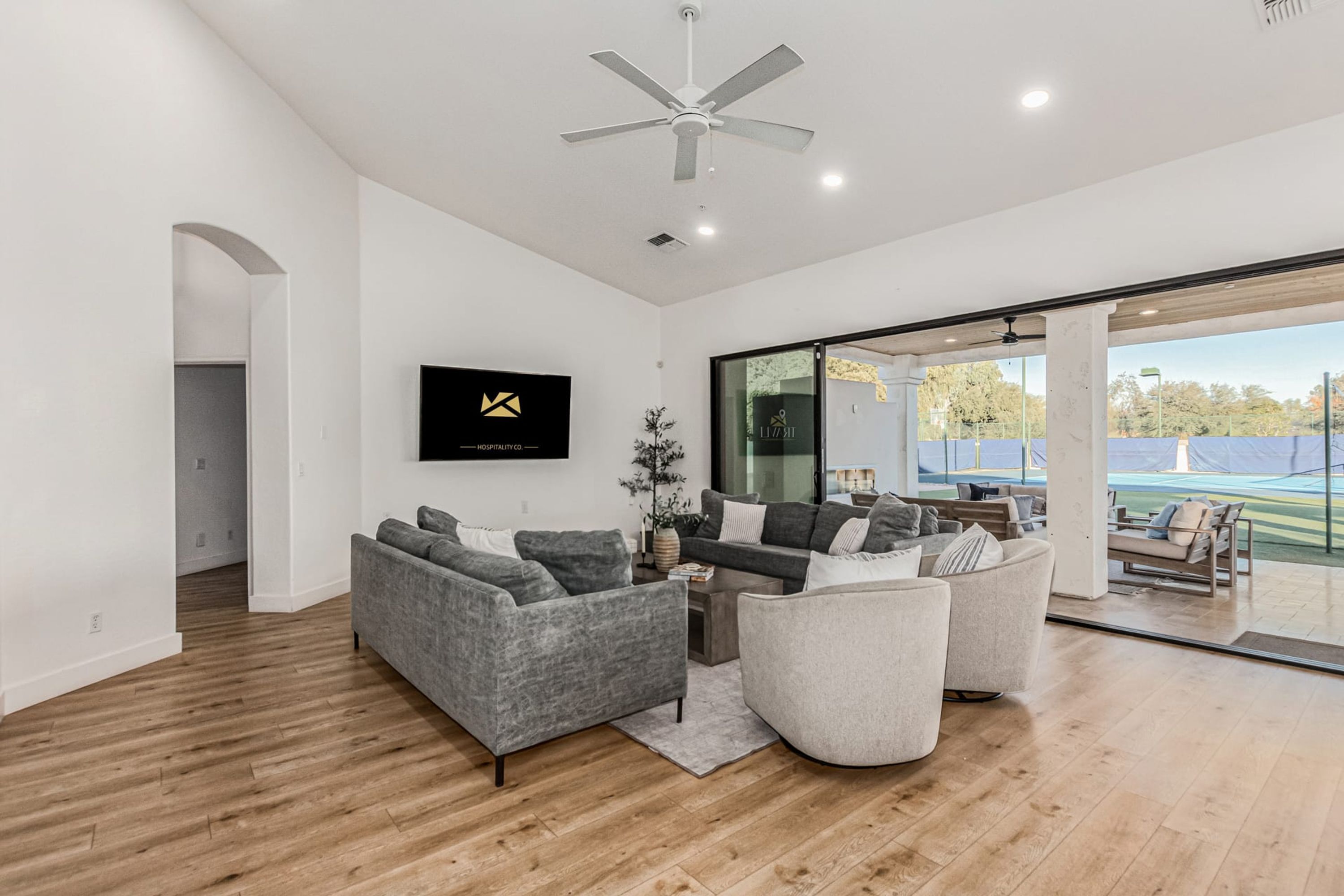 A modern living room with a grey sofa set, a flat-screen TV mounted on the wall, and a large glass door leading to an outdoor area.