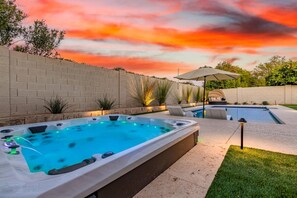Un bain à remous avec une piscine en arrière-plan, entouré d’un mur de briques et d’une zone herbeuse.