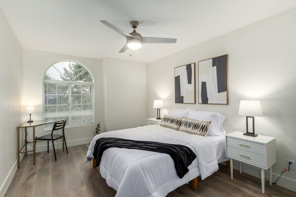 A bedroom with a bed, two bedside tables, a desk, a chair, a ceiling fan, and a window with blinds.