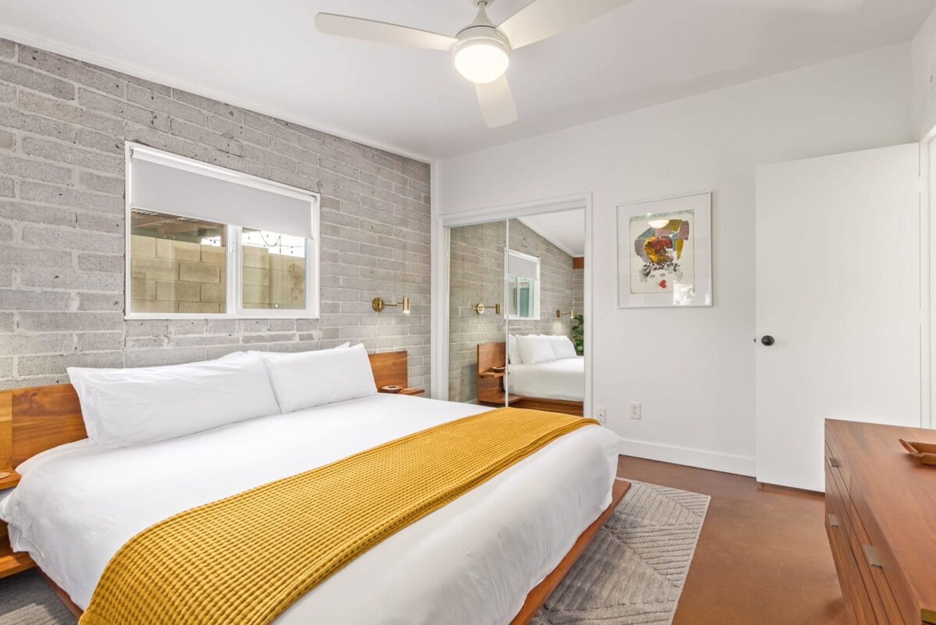 A bedroom with a large bed, a ceiling fan, a brick wall, a window, and a door.