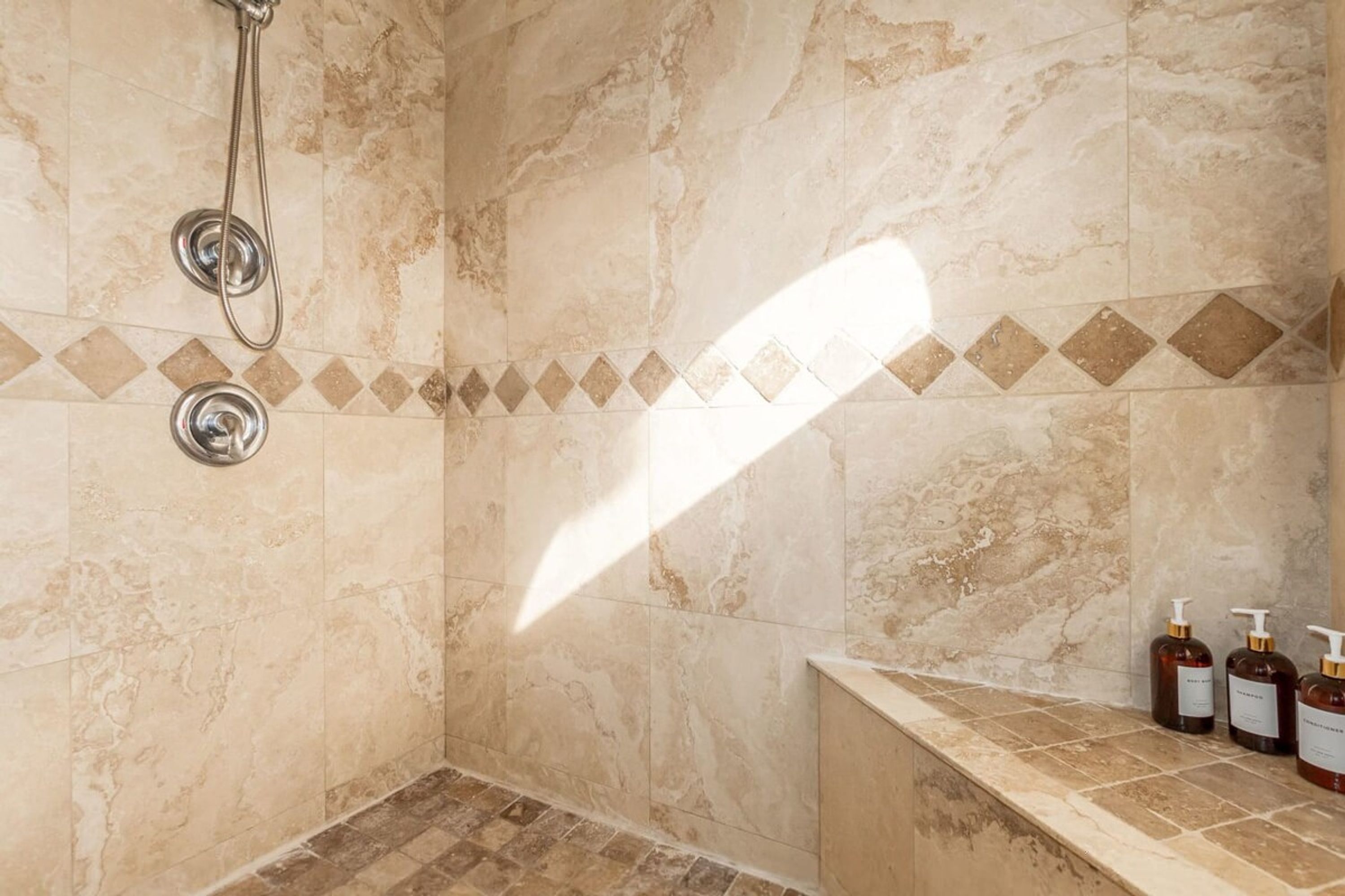 Ein Badezimmer mit beige farbenen Marmorfliesen, einer Dusche mit Duschbrausehandstück und einer Sitzbank mit drei Shampoo-Flaschen.