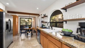 A modern kitchen with a stainless steel refrigerator, a dishwasher, and a coffee maker. There is a dining area with a table and chairs, and a window with a view of greenery. - Serenity Retreat Sedona Hot Tub Games Firepit (Sedona)