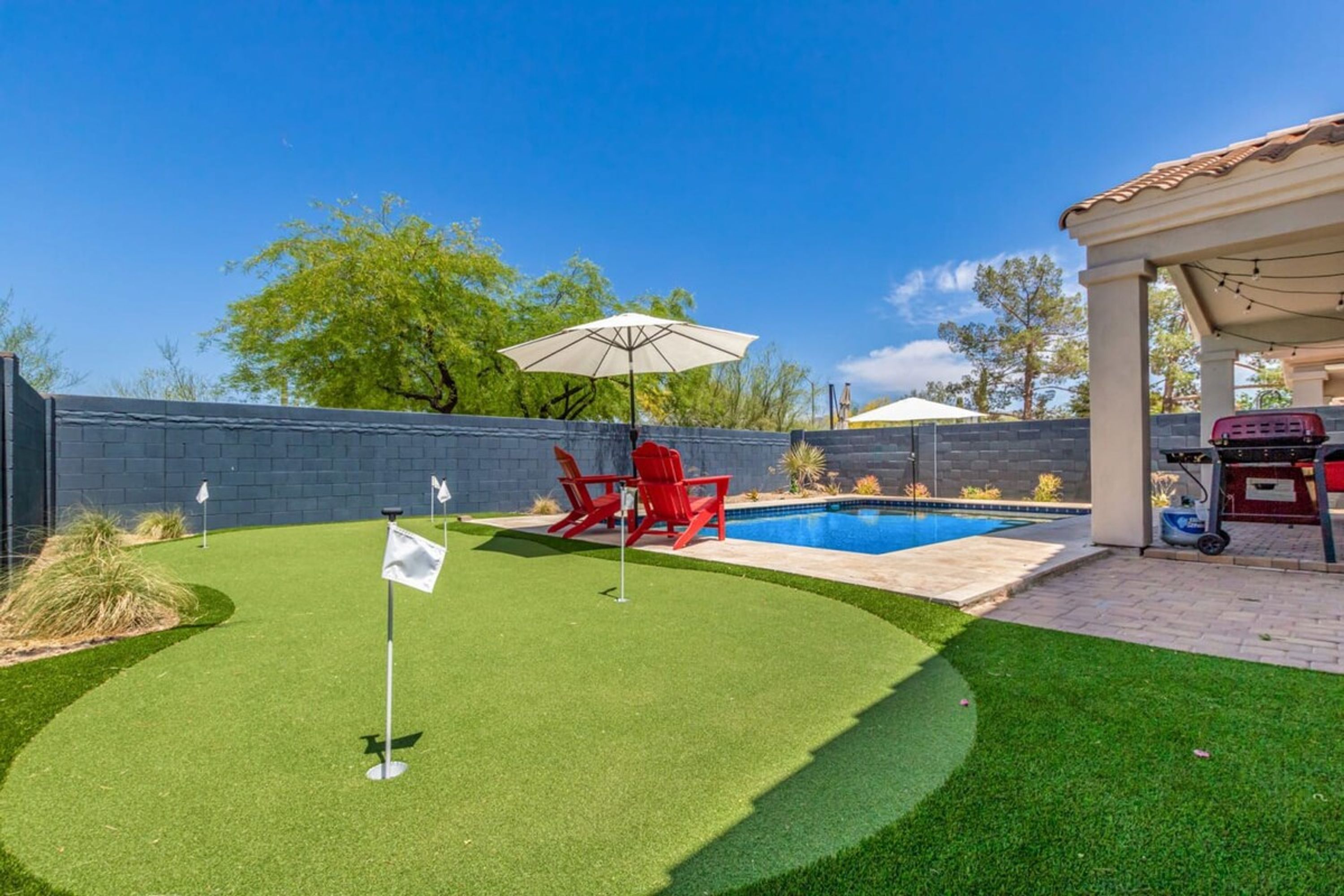 A backyard with a swimming pool, artificial grass, a red chair, and a barbecue grill.