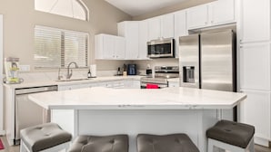 A modern kitchen with white cabinets, a large island with seating, stainless steel appliances, and a window with blinds. - Walk 2 TPC Mayo Swim Putt Grill Heated Pool (Scottsdale)