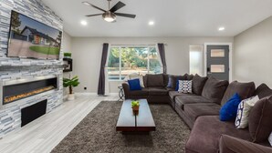 A modern living room with a stone fireplace, a ceiling fan, and a large window with curtains. - Kings Lux Escape Free Htd Pool Cabana BBQ Firepit (Scottsdale)