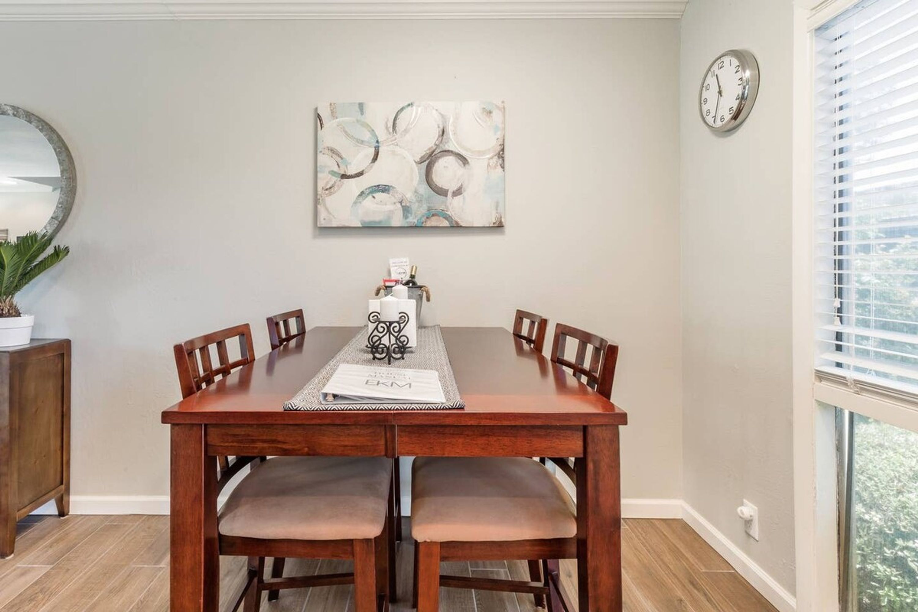 Un comedor con una mesa de madera, seis sillas, un espejo decorativo, un reloj y una ventana con persianas.