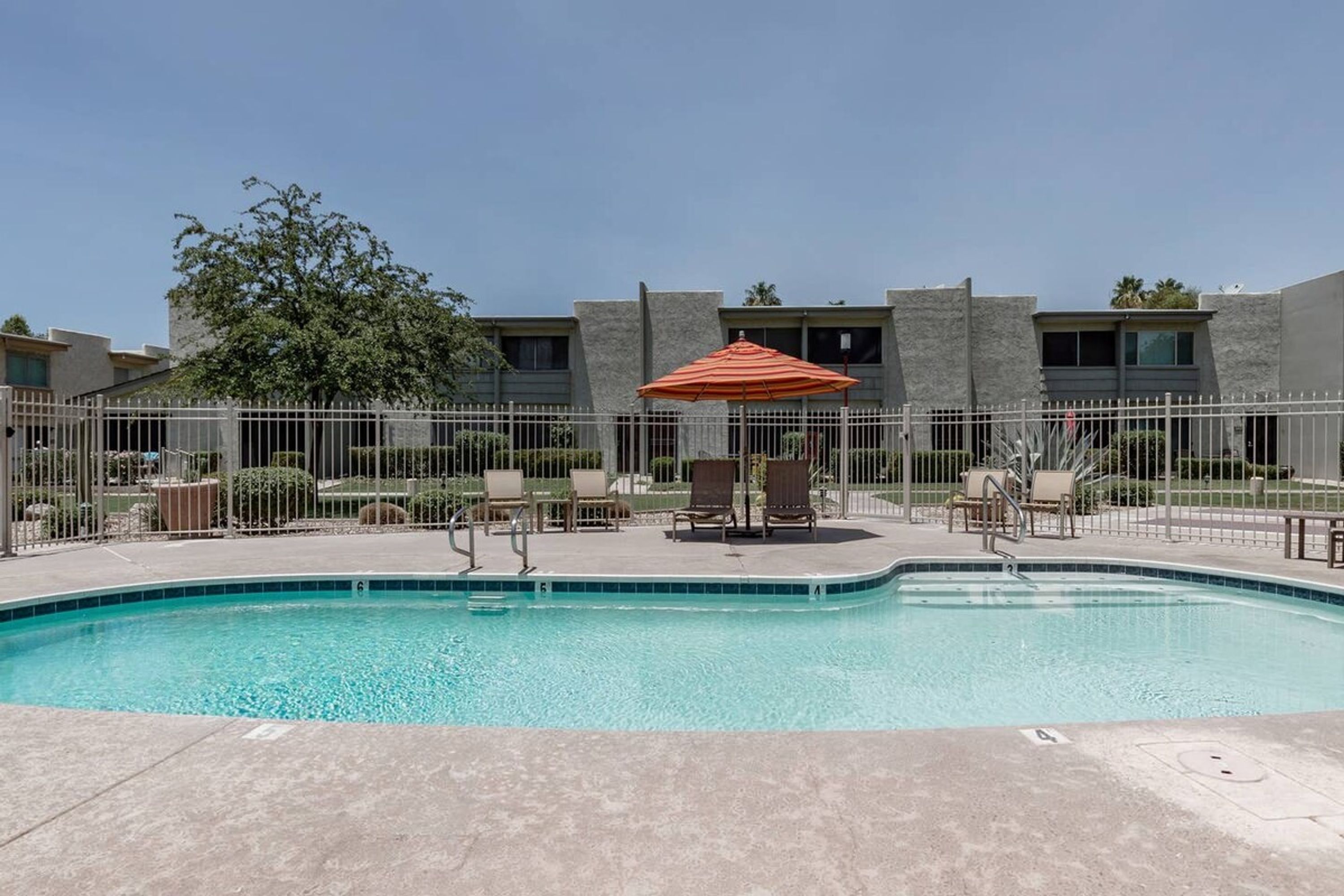 A swimming pool with lounge chairs and an umbrella, surrounded by a fence and buildings.