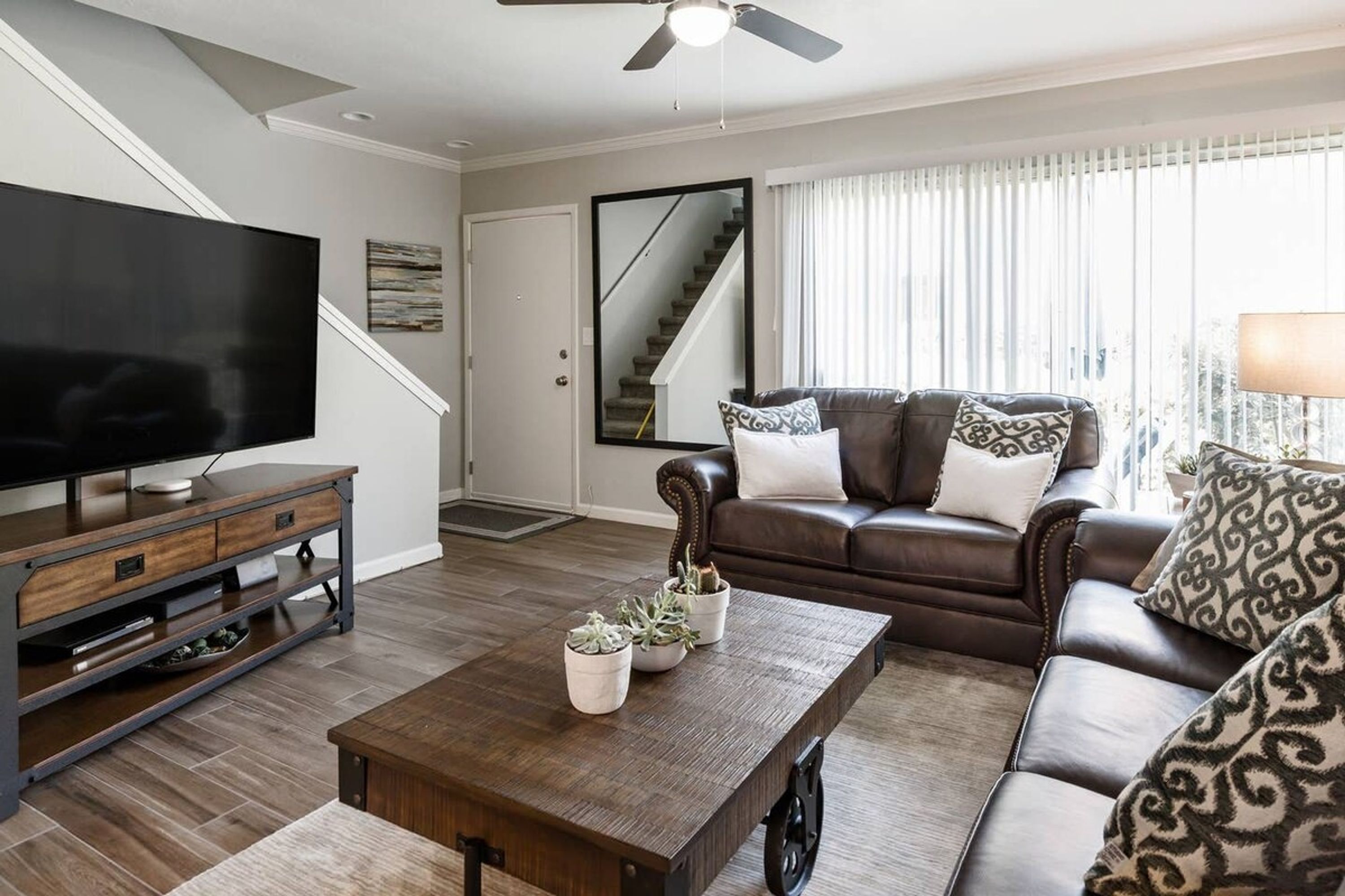 Een moderne woonkamer met een bruine leren bank, een houten salontafel met planten, een flatscreen televisie op een houten standaard en een plafondventilator.