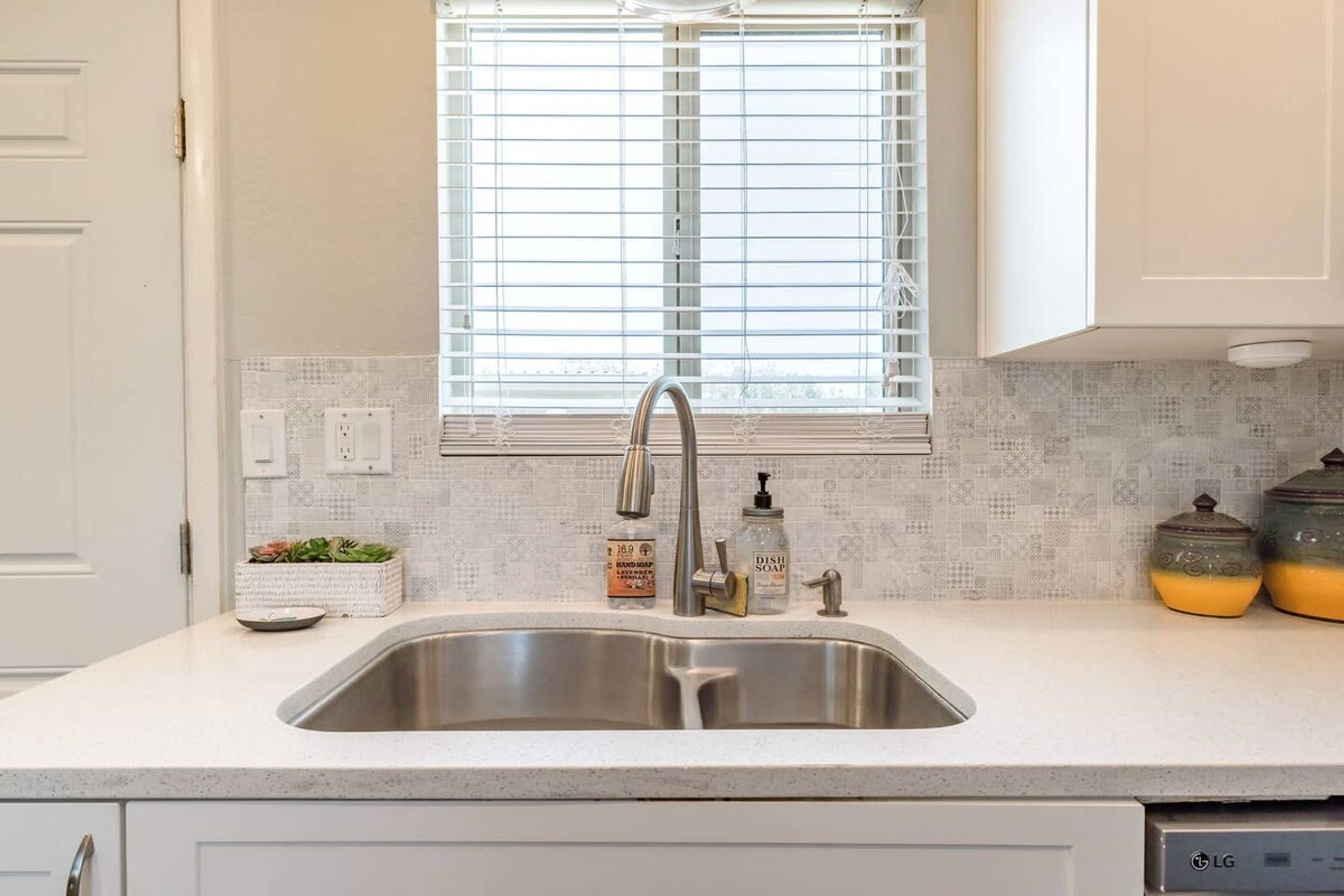 A kitchen with a stainless steel sink, white cabinets, a window with blinds, and a backsplash with a circuit board pattern.