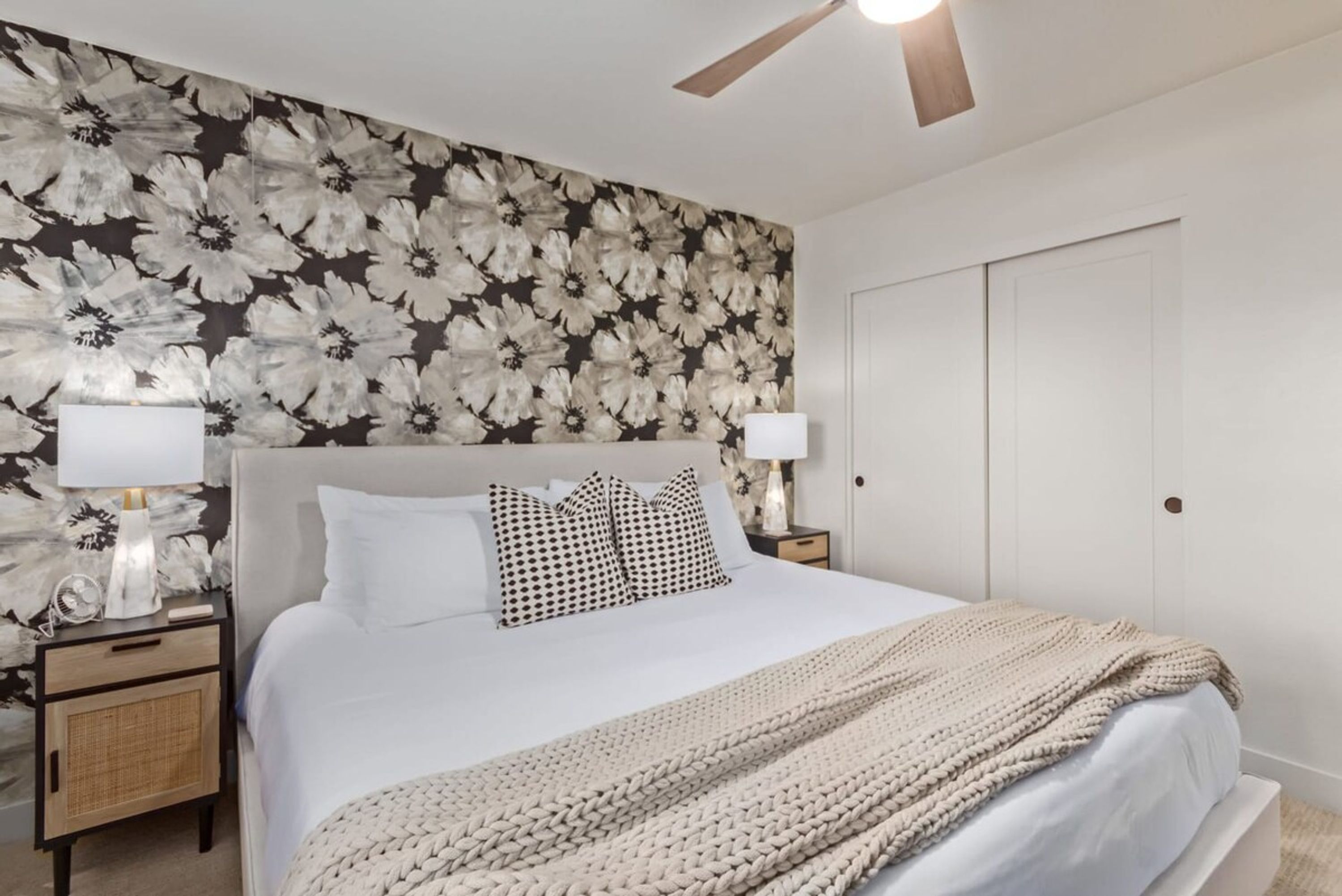 A bedroom with a large bed, a ceiling fan, a nightstand with a lamp, and a floral wallpaper.