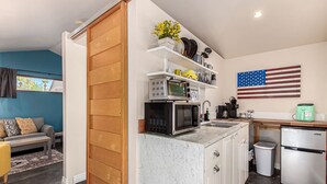 A compact kitchen with a microwave, sink, and refrigerator. There is a wooden cabinet and a shelf with kitchenware. An American flag is displayed on the wall. - Coronado Casita - Mins to DT Airport Hospitals (Phoenix)