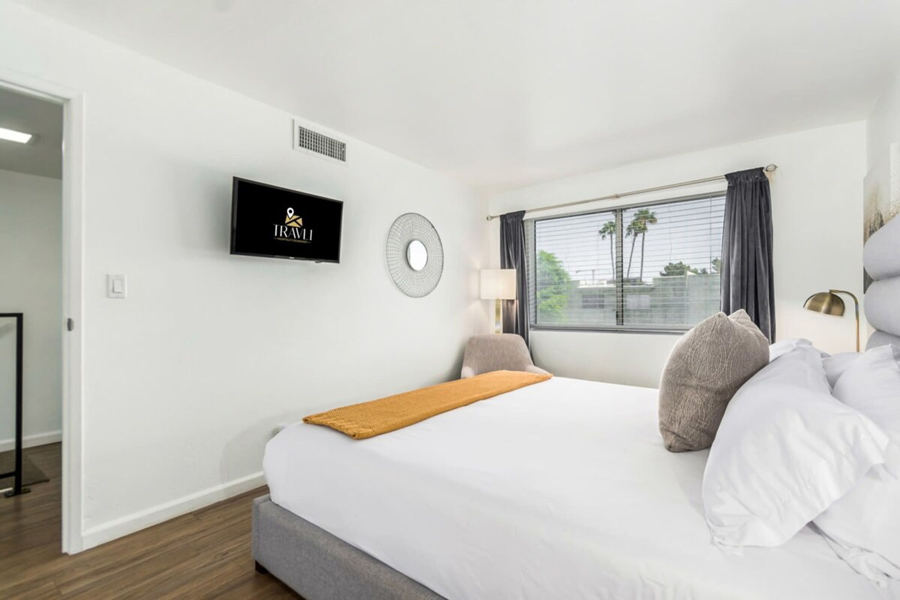 A hotel room with a bed, a TV mounted on the wall, a window with curtains, and a view of palm trees outside.