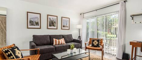 A modern living room with a grey sofa, a wooden armchair, a glass coffee table, and a patterned rug. There are three framed pictures on the wall, a lamp, and a large window with curtains.