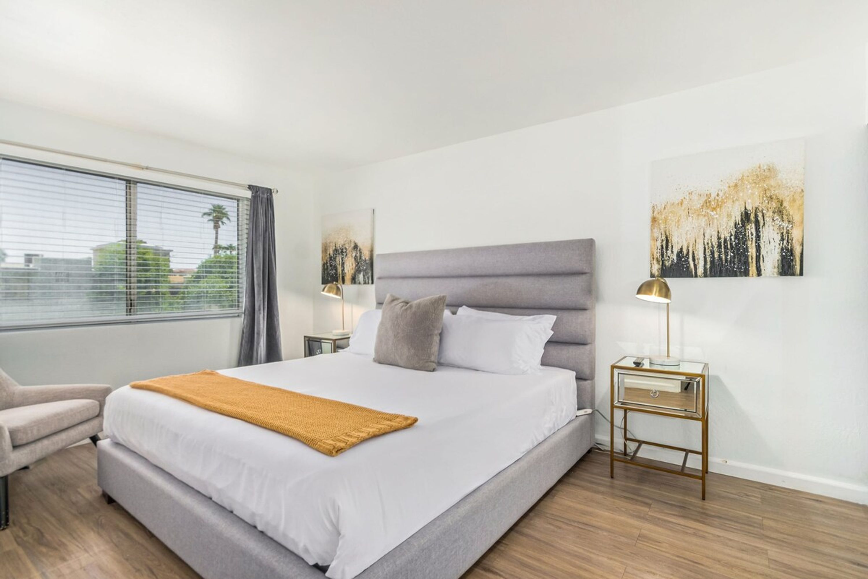 A modern bedroom with a large bed, a bedside table with a lamp, and a view of the outdoors through a window.