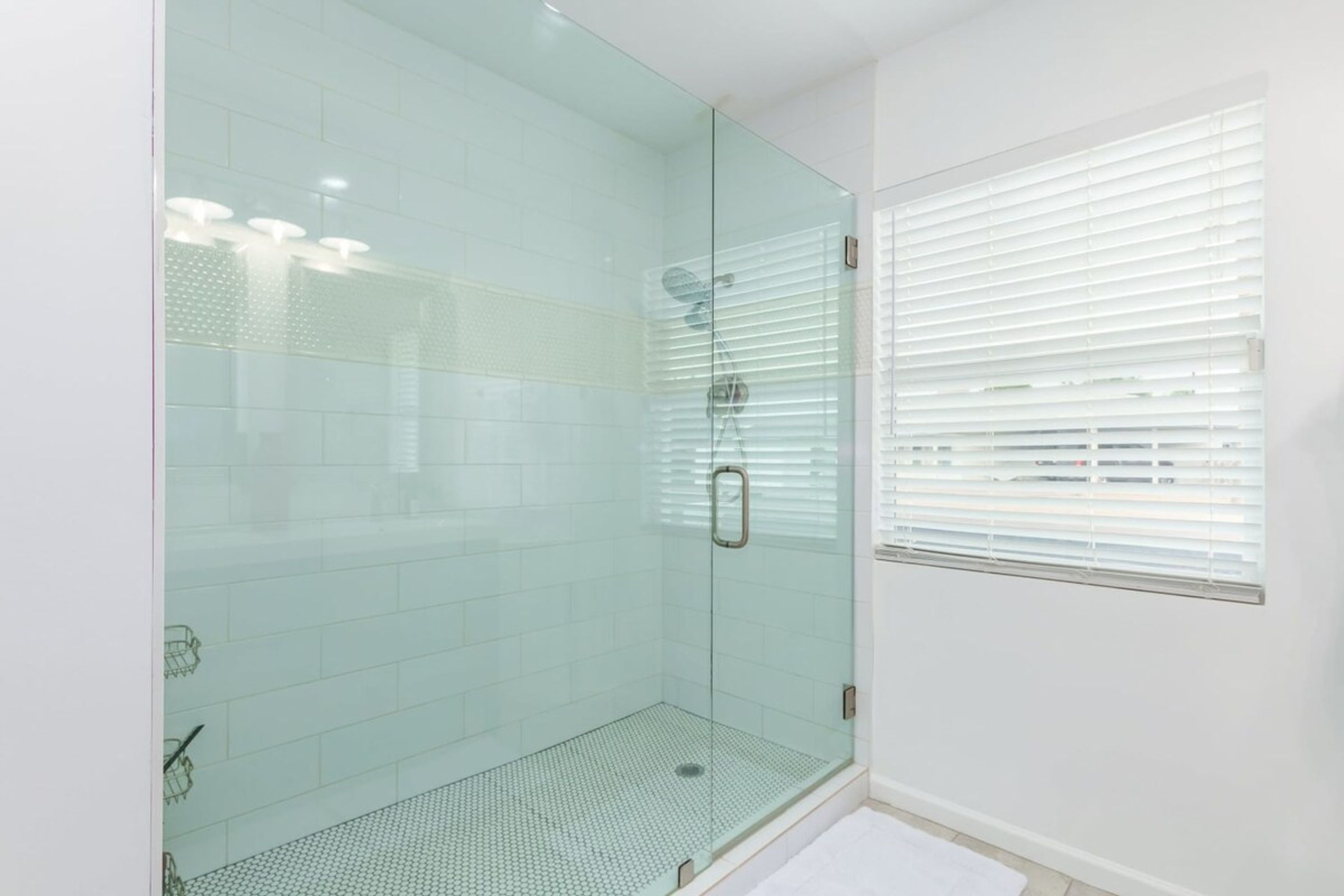 A modern bathroom with a glass shower enclosure, white tiled walls, and a window with blinds.
