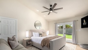 Une chambre avec un lit, des tables de chevet, un ventilateur de plafond, un grand miroir et une vue sur l’extérieur, accessible par une porte-fenêtre coulissante.