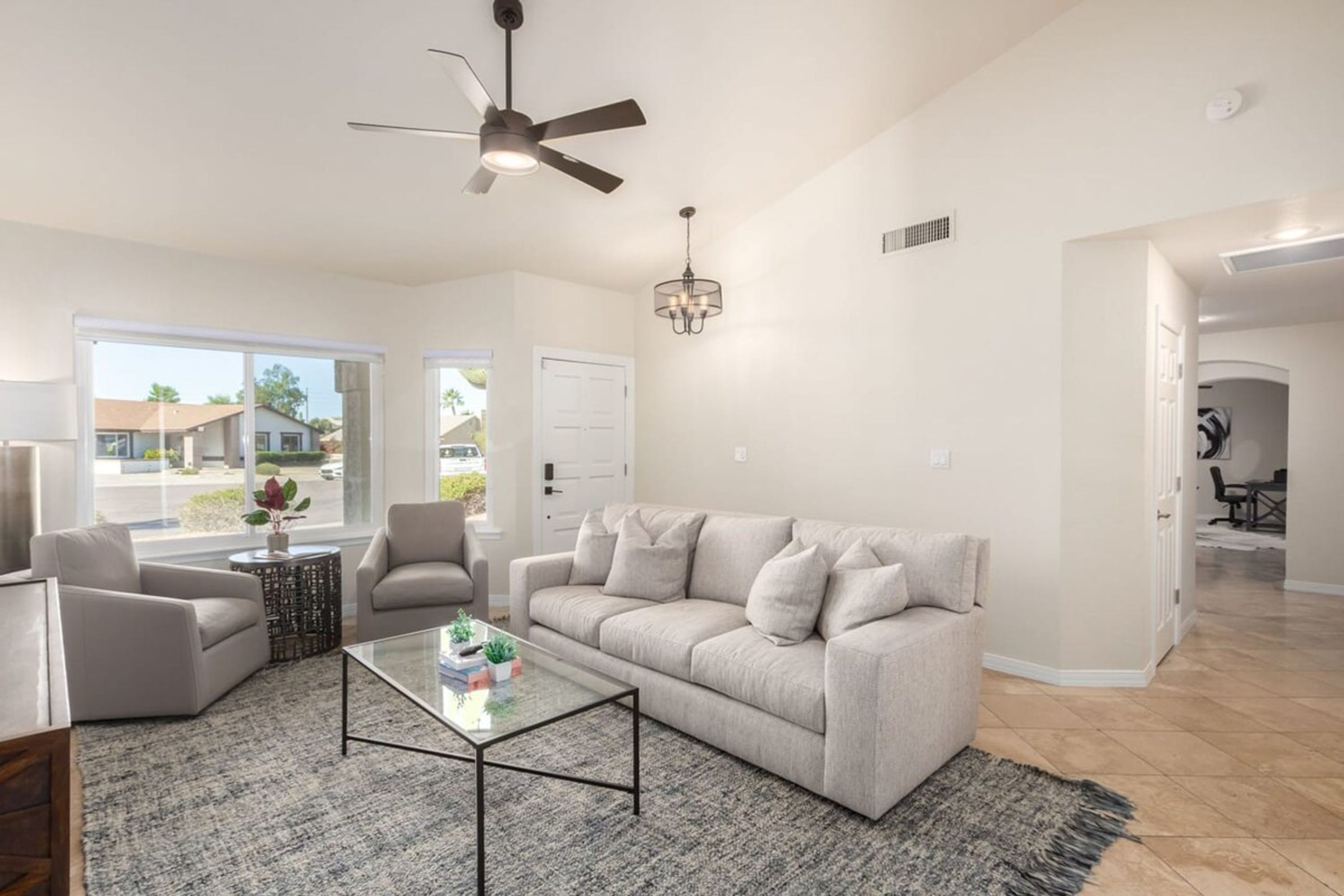 A spacious living room with a large window, a ceiling fan, a chandelier, and a modern coffee table.