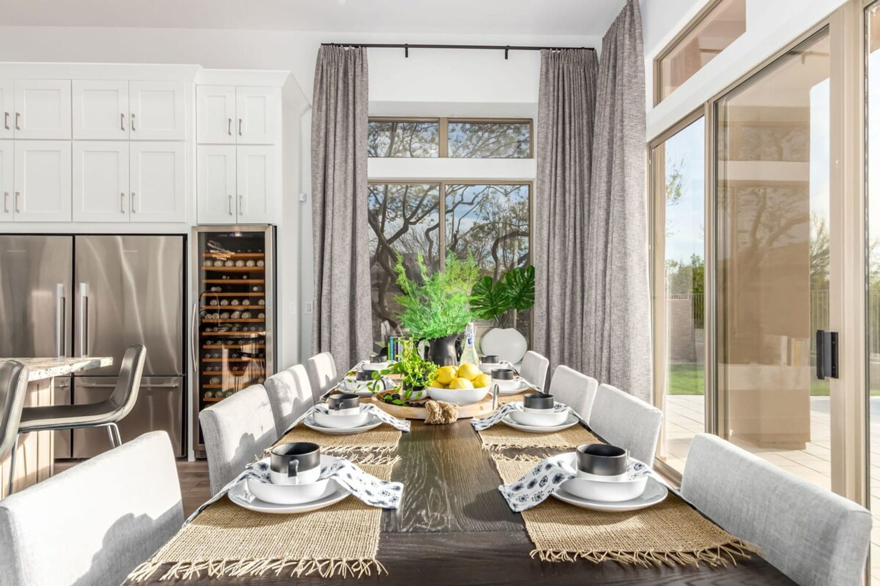A dining area with a long table set for six, surrounded by white cabinets and a large window with curtains.
