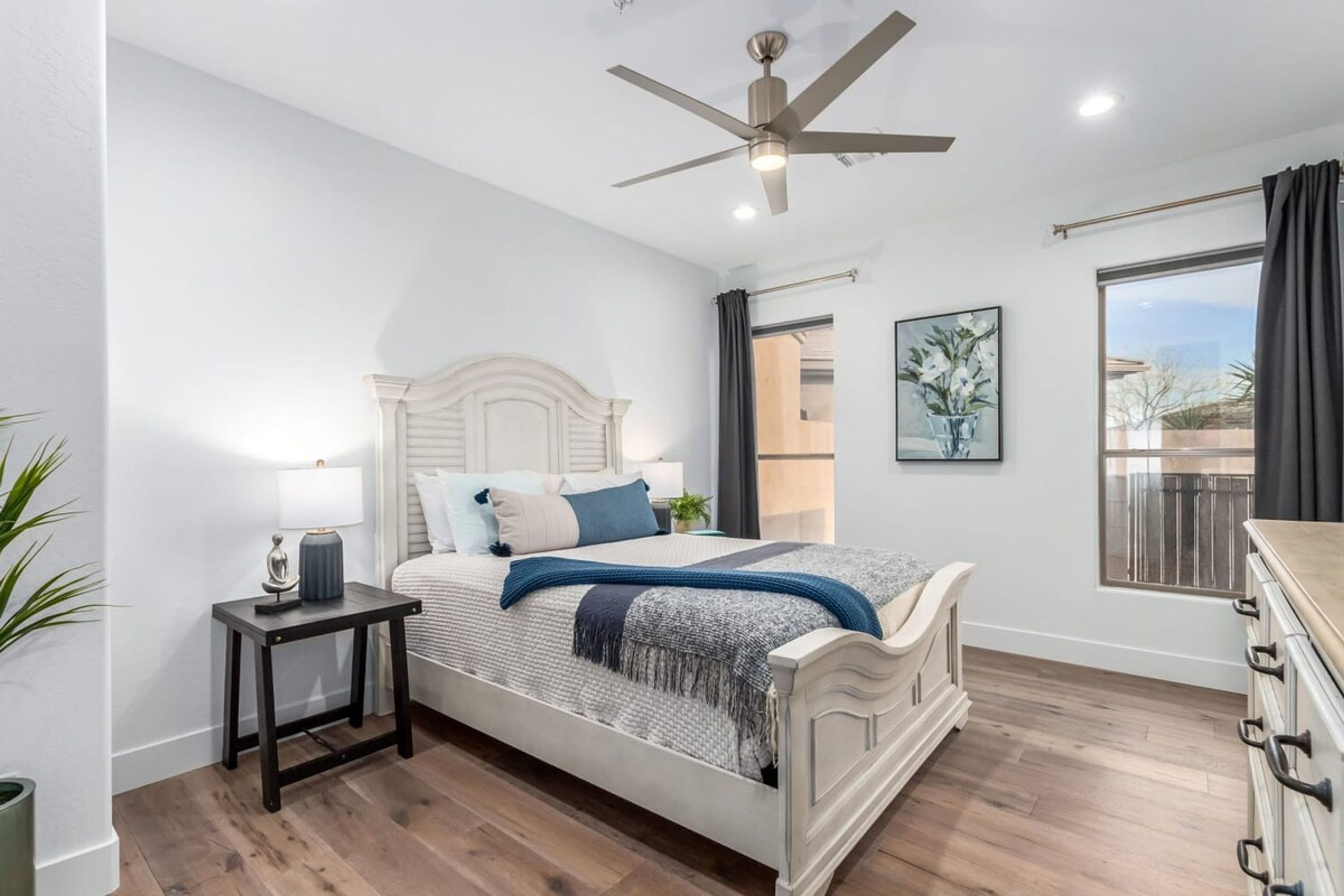 A bedroom with a bed, a nightstand, a ceiling fan, a window with curtains, and a plant.