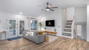 Ein modernes Wohnzimmer mit grauem Sofa, Holzboden, Deckenventilator, Flachbildfernseher und einer Treppe.