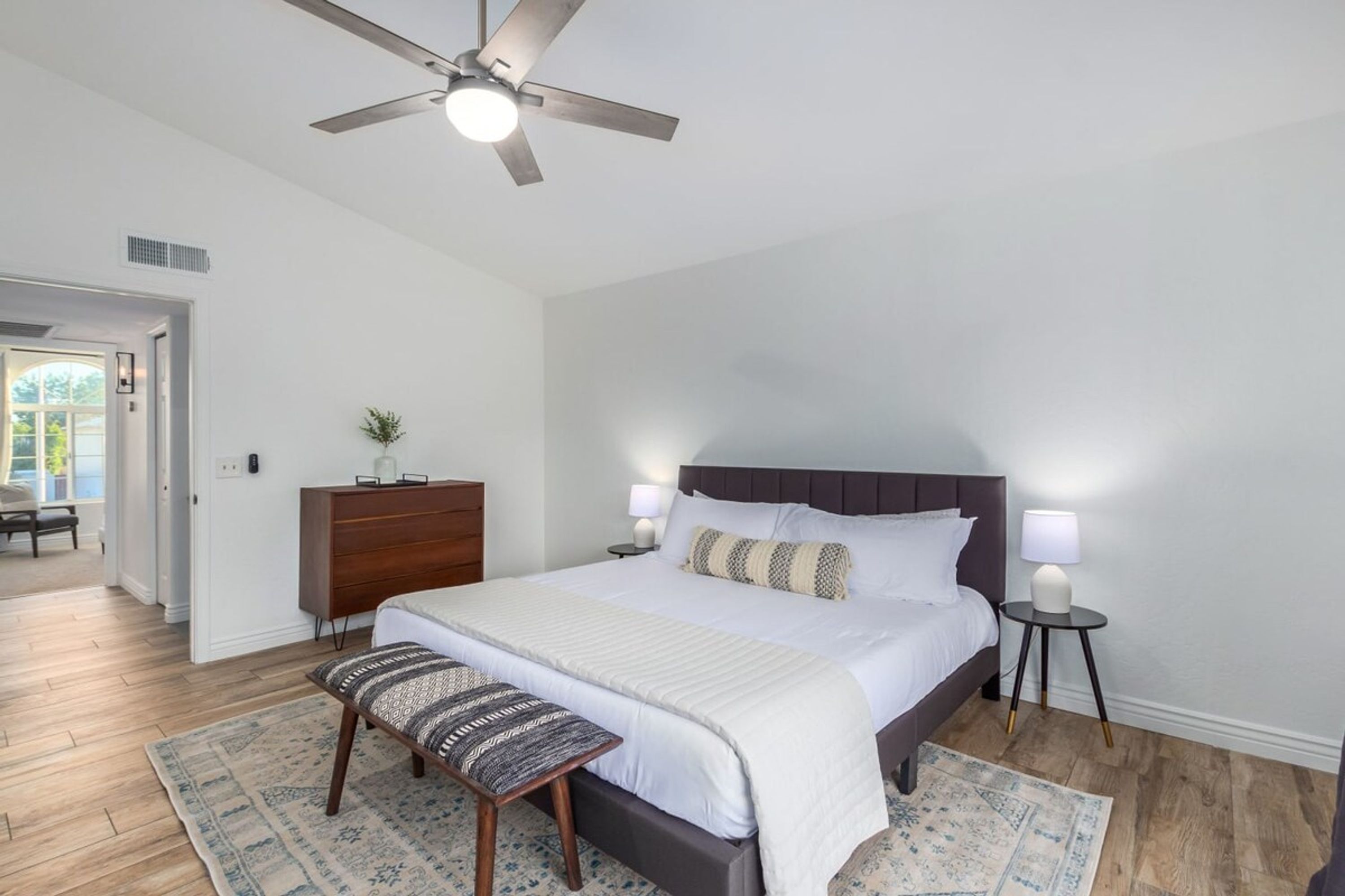 A bedroom with a large bed, a ceiling fan, a nightstand, a dresser, and a bench.
