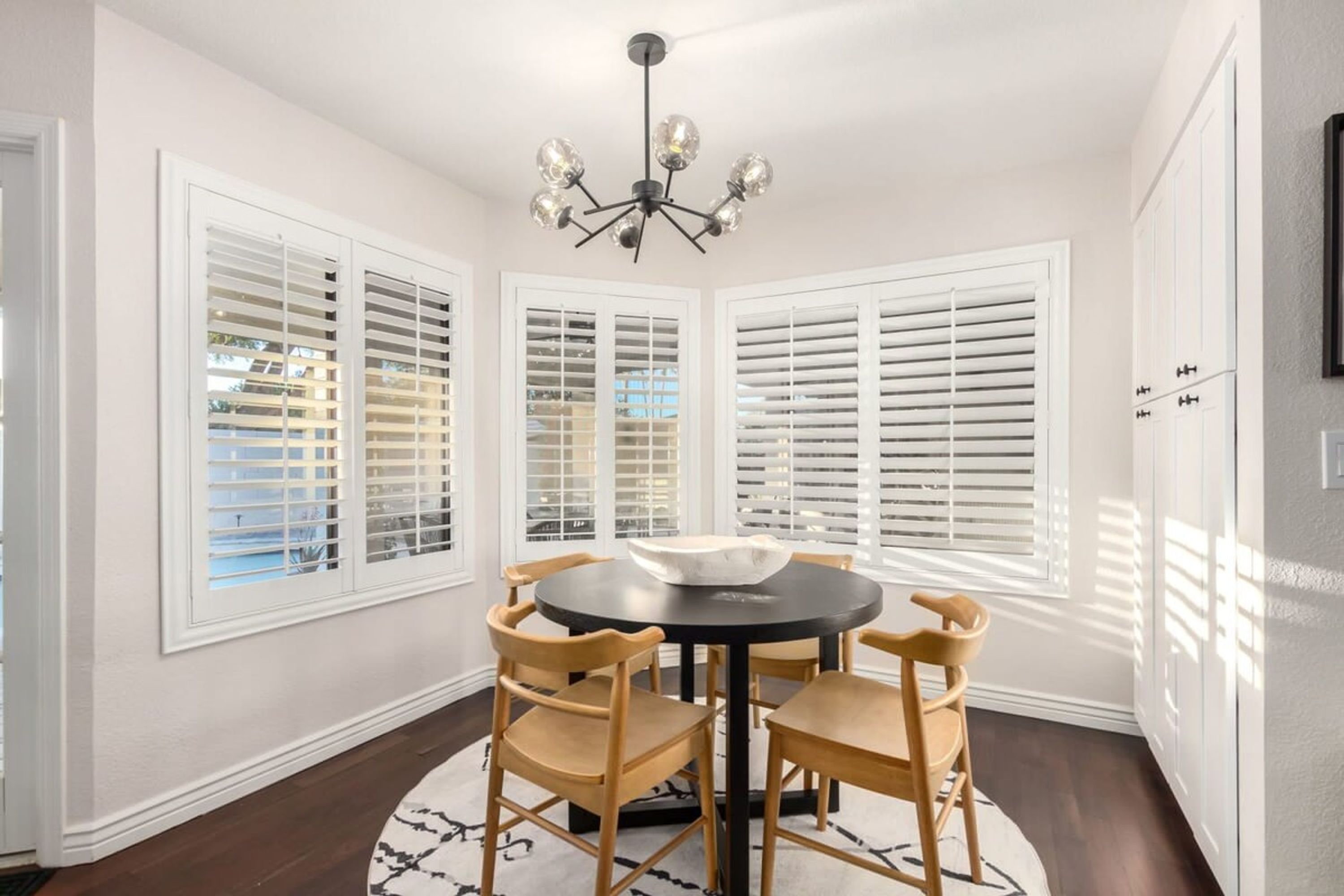 A dining area with a round table and four chairs, a chandelier, and large windows with blinds.