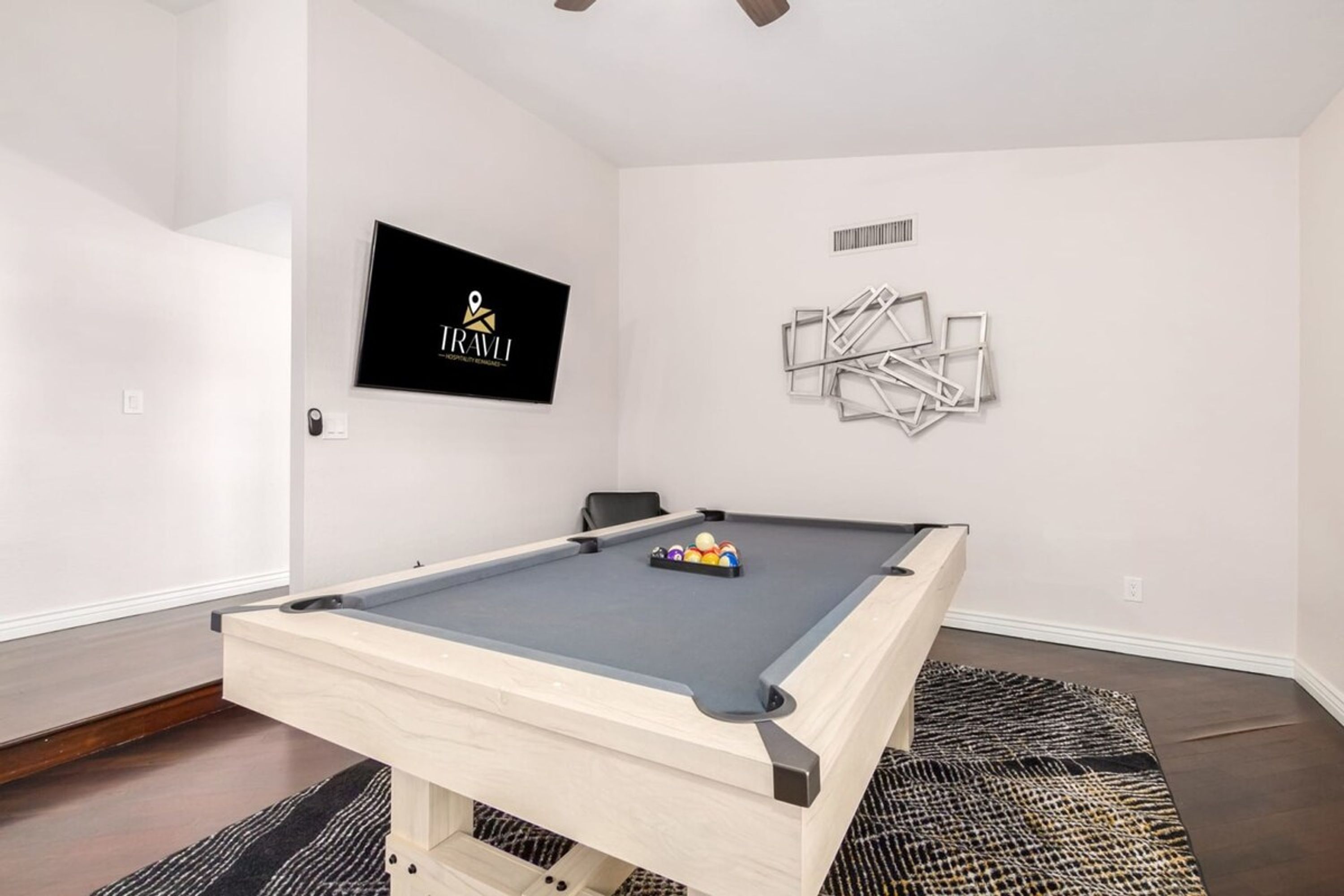 A pool table in a room with white walls, a mounted television, and a decorative wall piece.